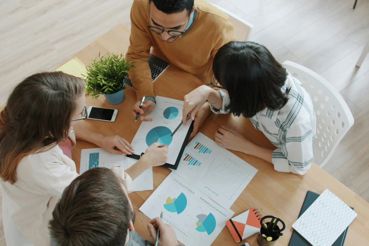 A group of four people sit around a wooden table, discussing charts and graphs.
