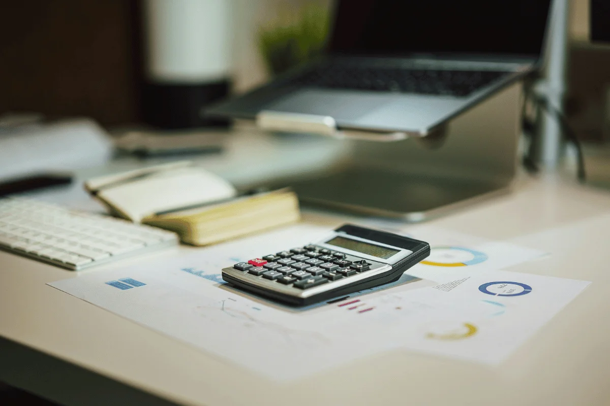Calculator on a desk with financial charts, open notebook, keyboard, and laptop.