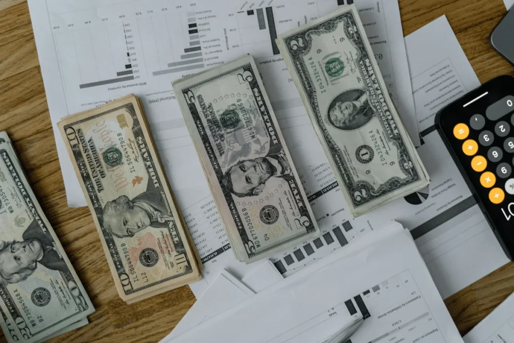 Three US dollar bills (five, ten, and twenty) placed on financial documents and a calculator on a wooden table.