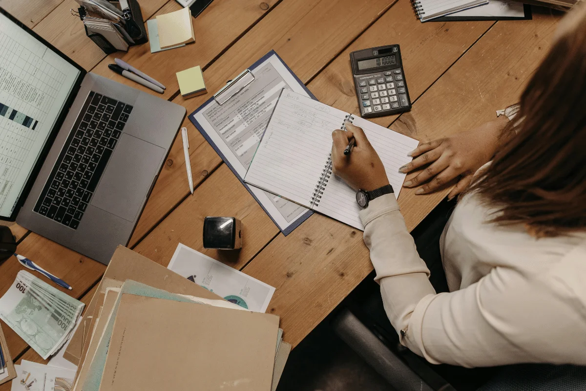 A person writes in a notebook at a wooden desk, surrounded by a laptop, calculator, stationery, and papers.