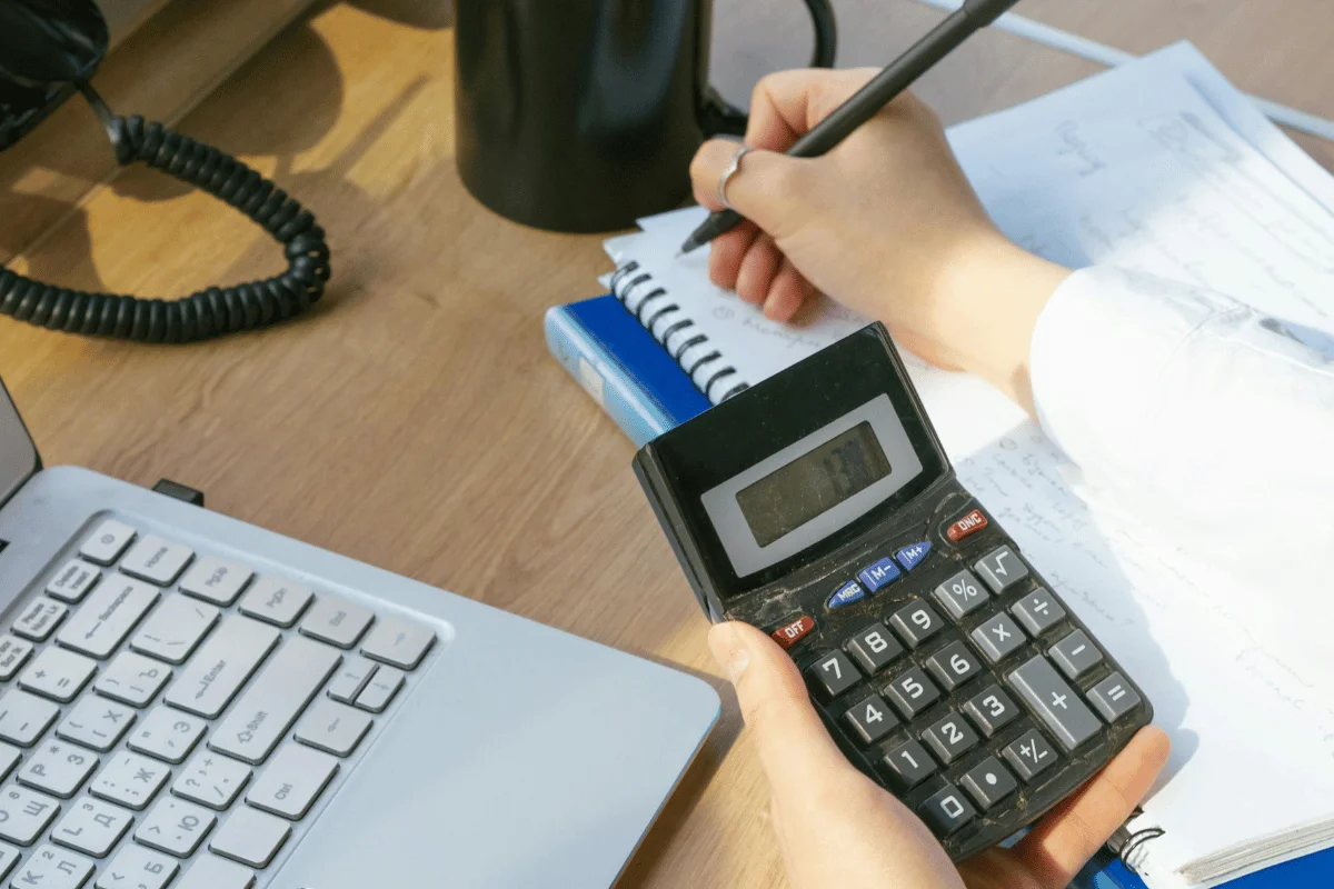 A person writes in a spiral notebook while holding a calculator.