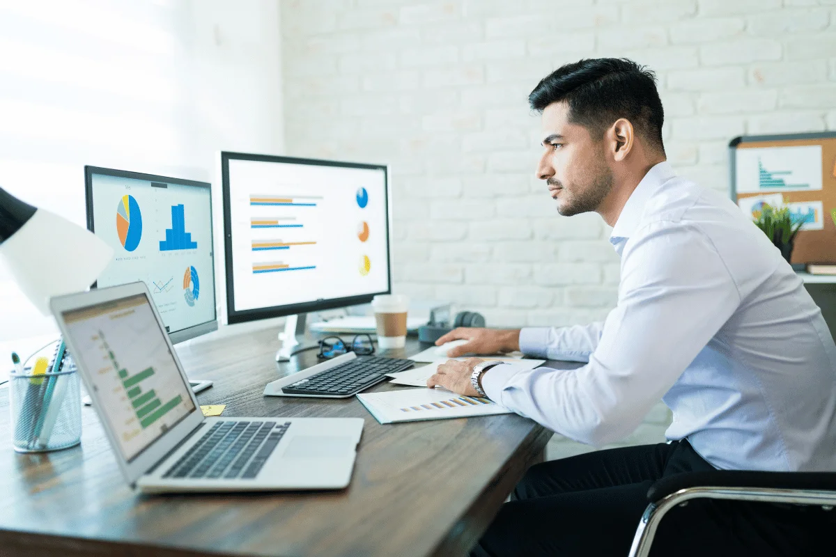 A man in a white shirt works at a wooden desk with a laptop and two monitors displaying colorful charts, conveying focus and productivity.