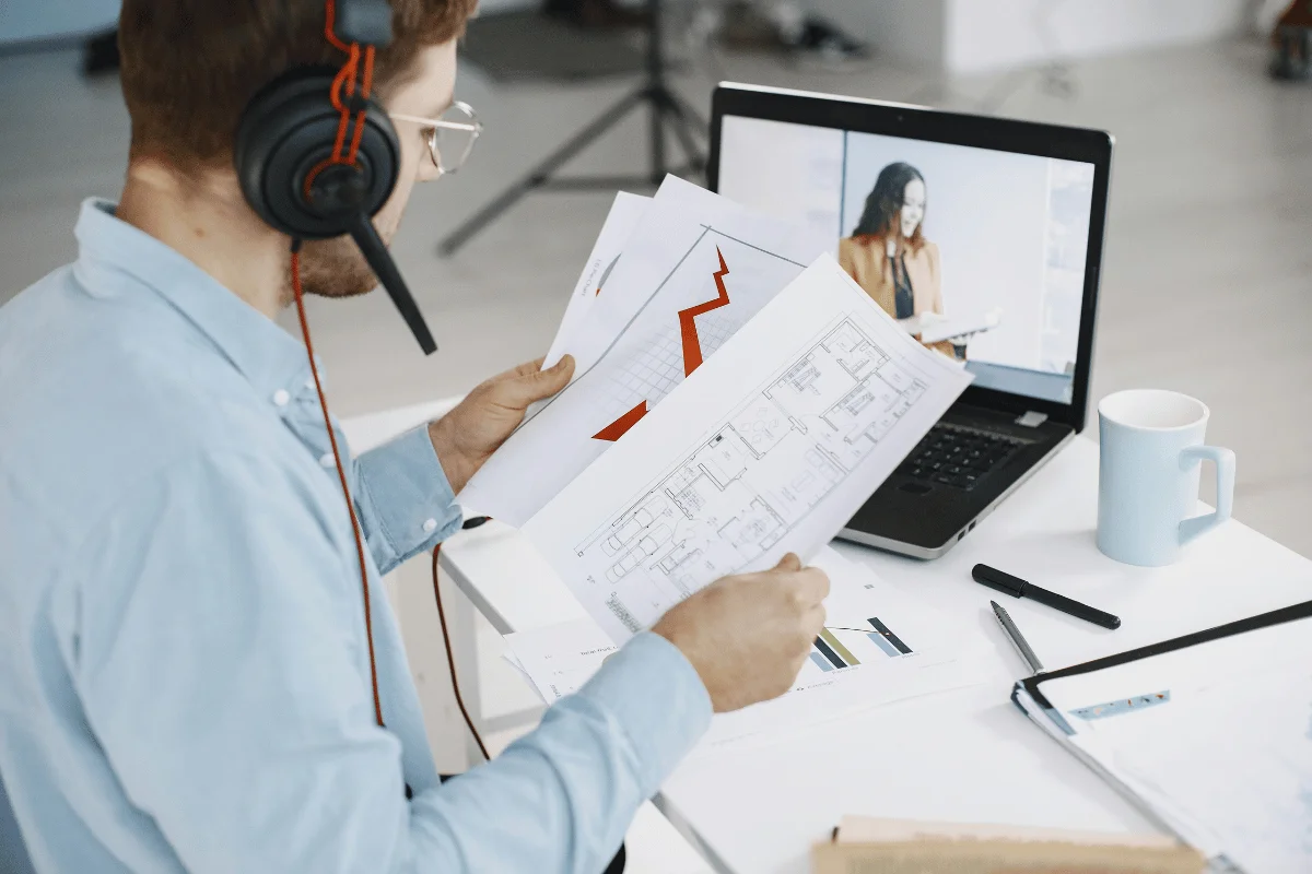 Man with headphones reviews graphs and floor plans at desk, while video conferencing on laptop.