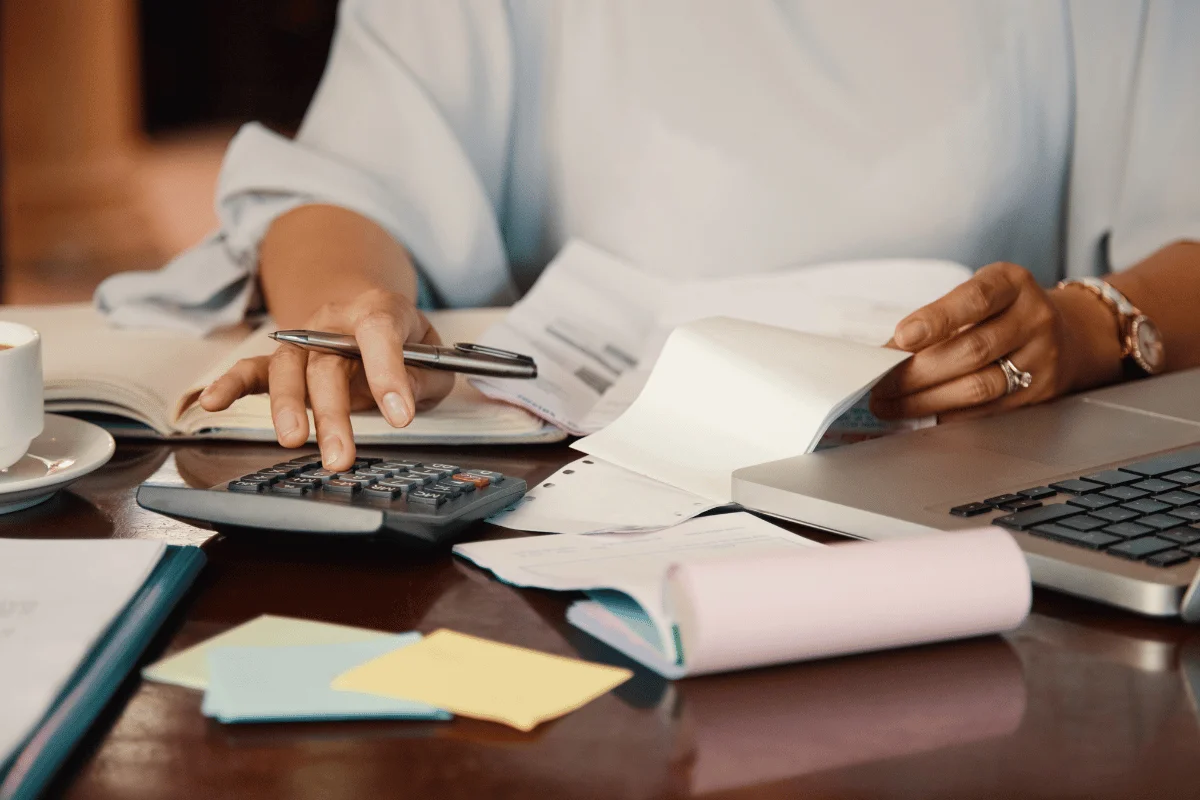 Hands managing finances with a calculator, pen, and receipts on a table, alongside a laptop and stacked papers. The scene conveys focus and organization.
